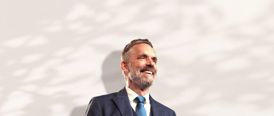 Jordan Peterson lächelnd im dunkelblauen Anzug vor heller Wand mit Blattschatten