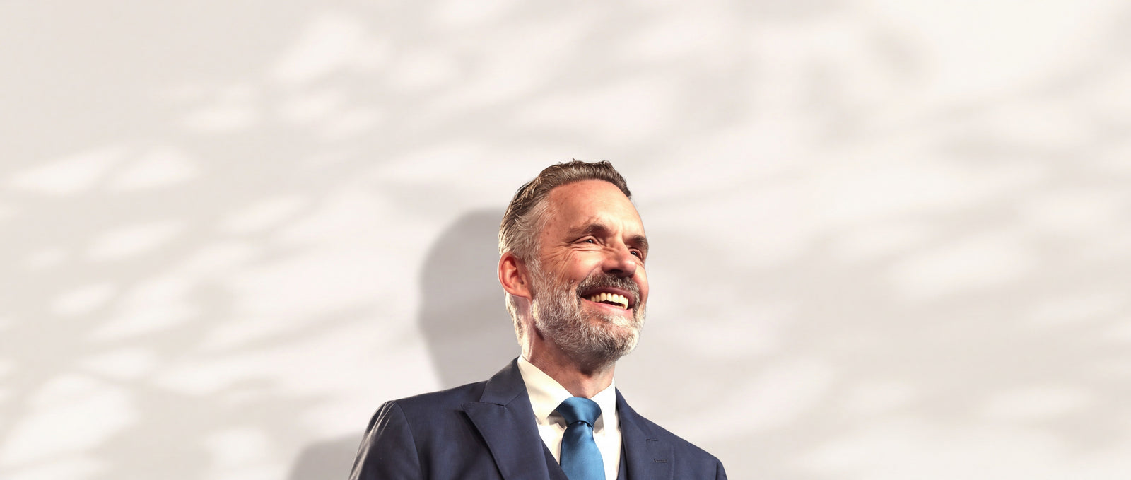 Jordan Peterson lächelnd im dunkelblauen Anzug vor heller Wand mit Blattschatten
