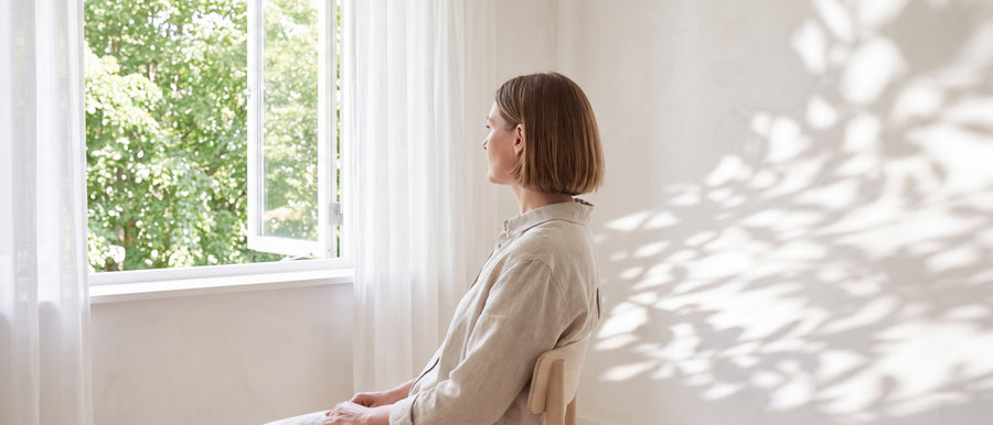 Frau sitzt ruhig am offenen Fenster und schaut ins Grüne, weiche Blattschatten an der hellen Wand